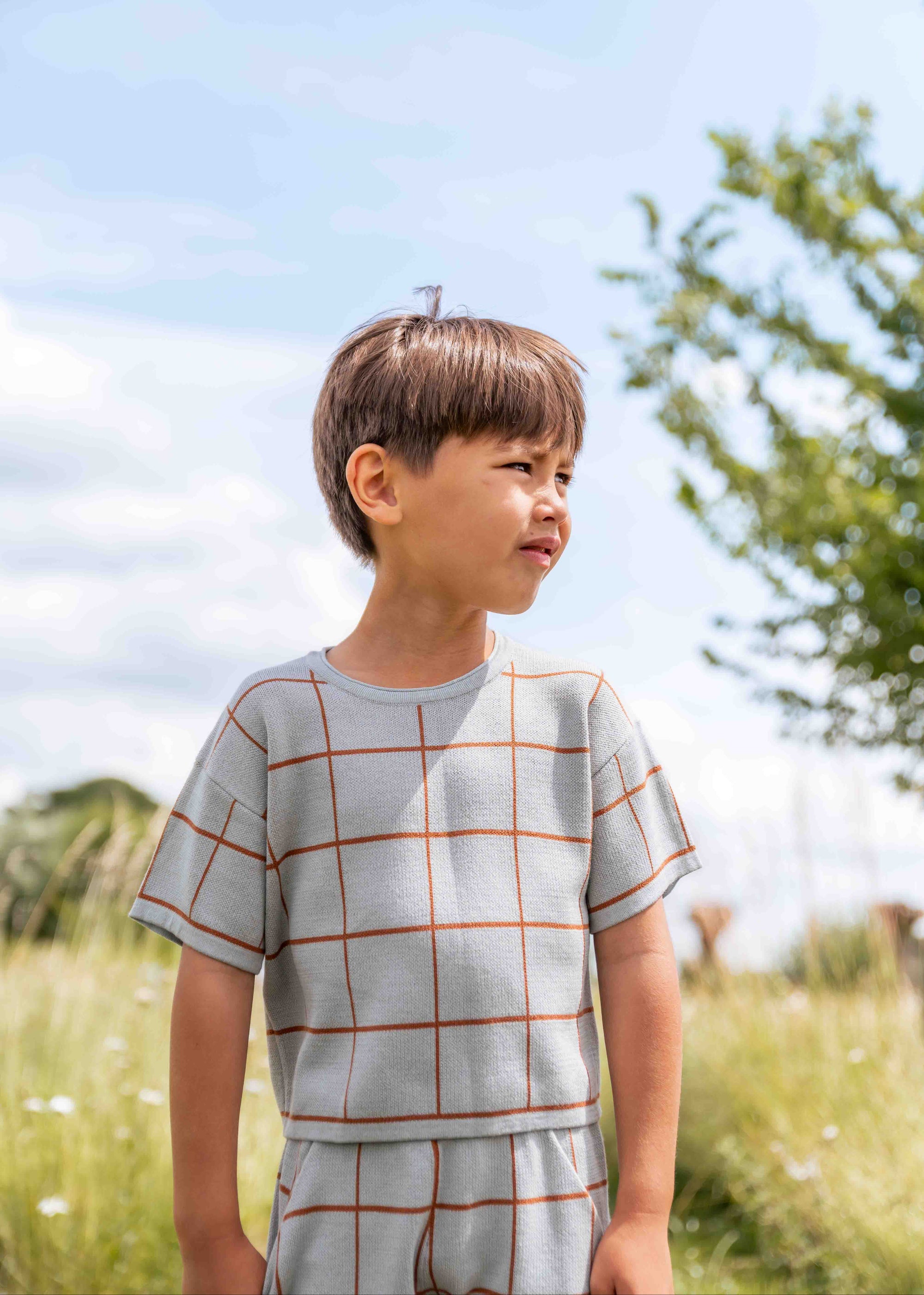 Child wearing a checkered outfit standing in a grassy field with a blue sky.