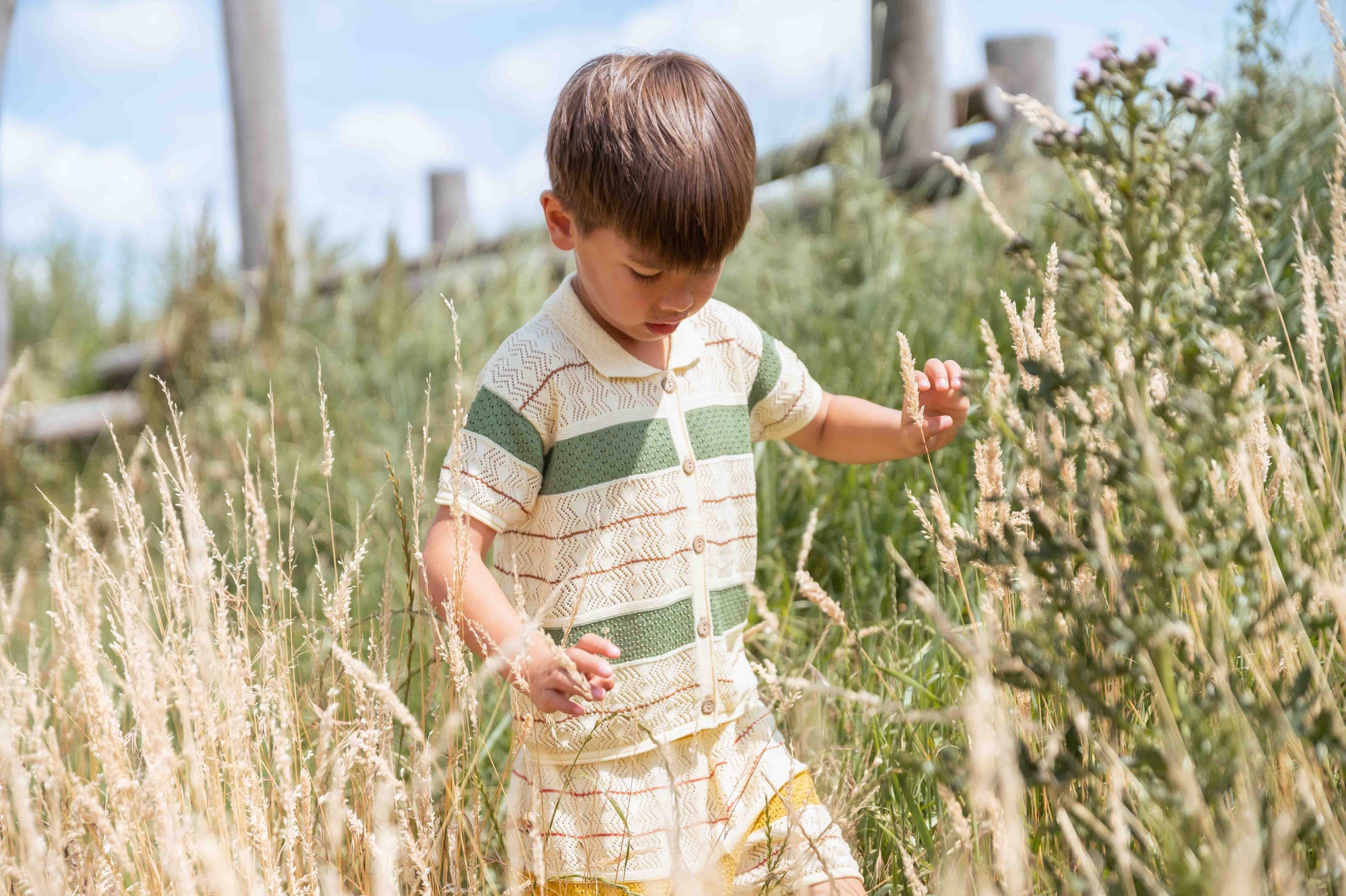 Child in a striped shirt standing in tall grass with a wooden fence in the background