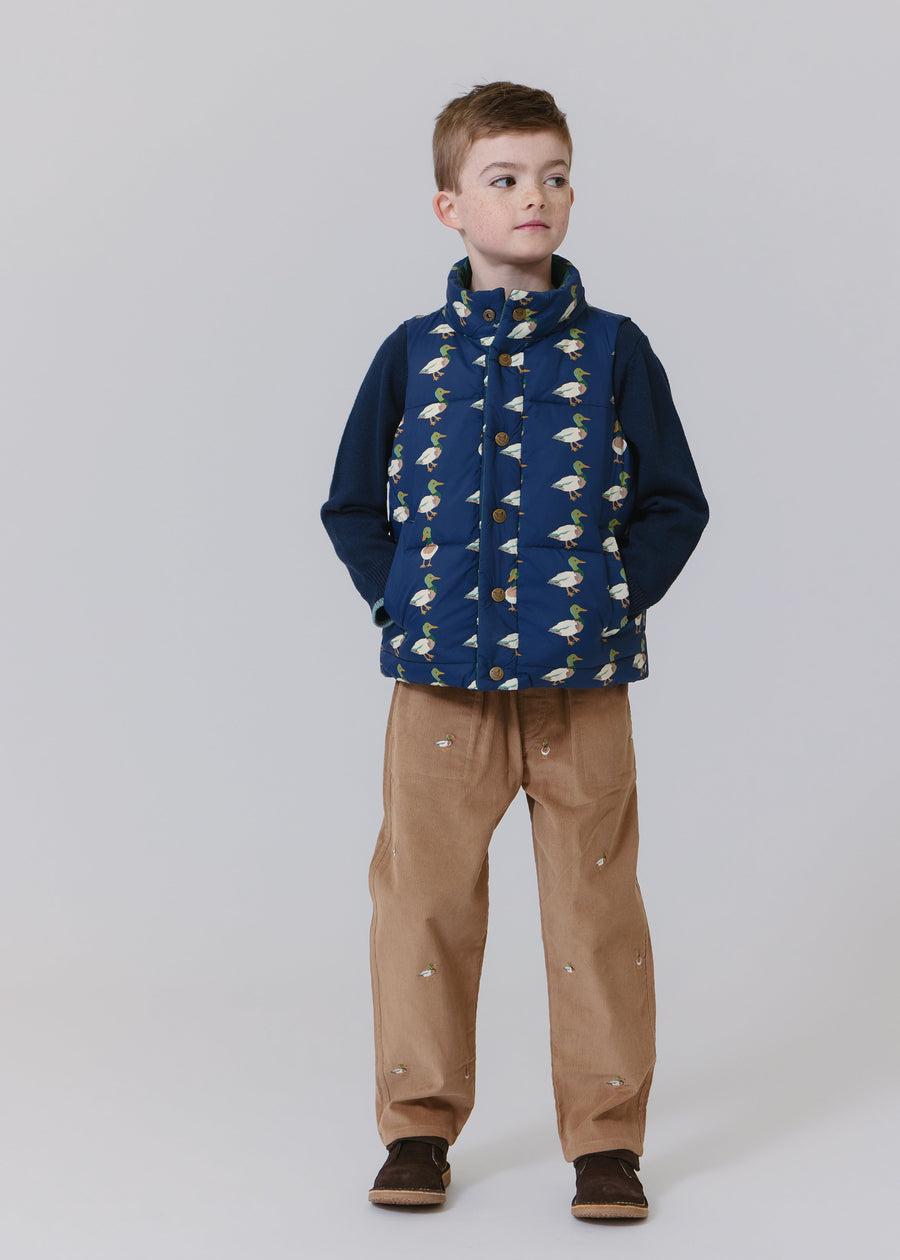 Child wearing a blue patterned vest over a dark sweater with brown pants on a white background