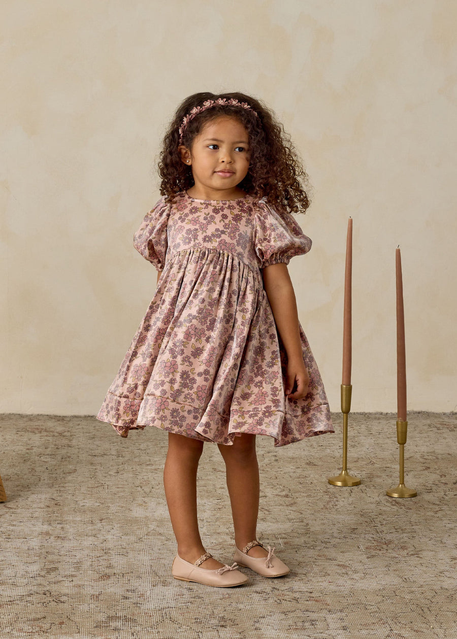 Young girl in a floral dress standing in a room with candles and a vase.