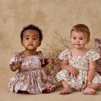 Two young children in floral dresses sitting on a beige surface with flowers in the background.