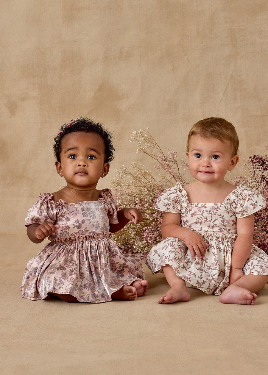 Two young children in floral dresses sitting on a beige surface with flowers in the background.