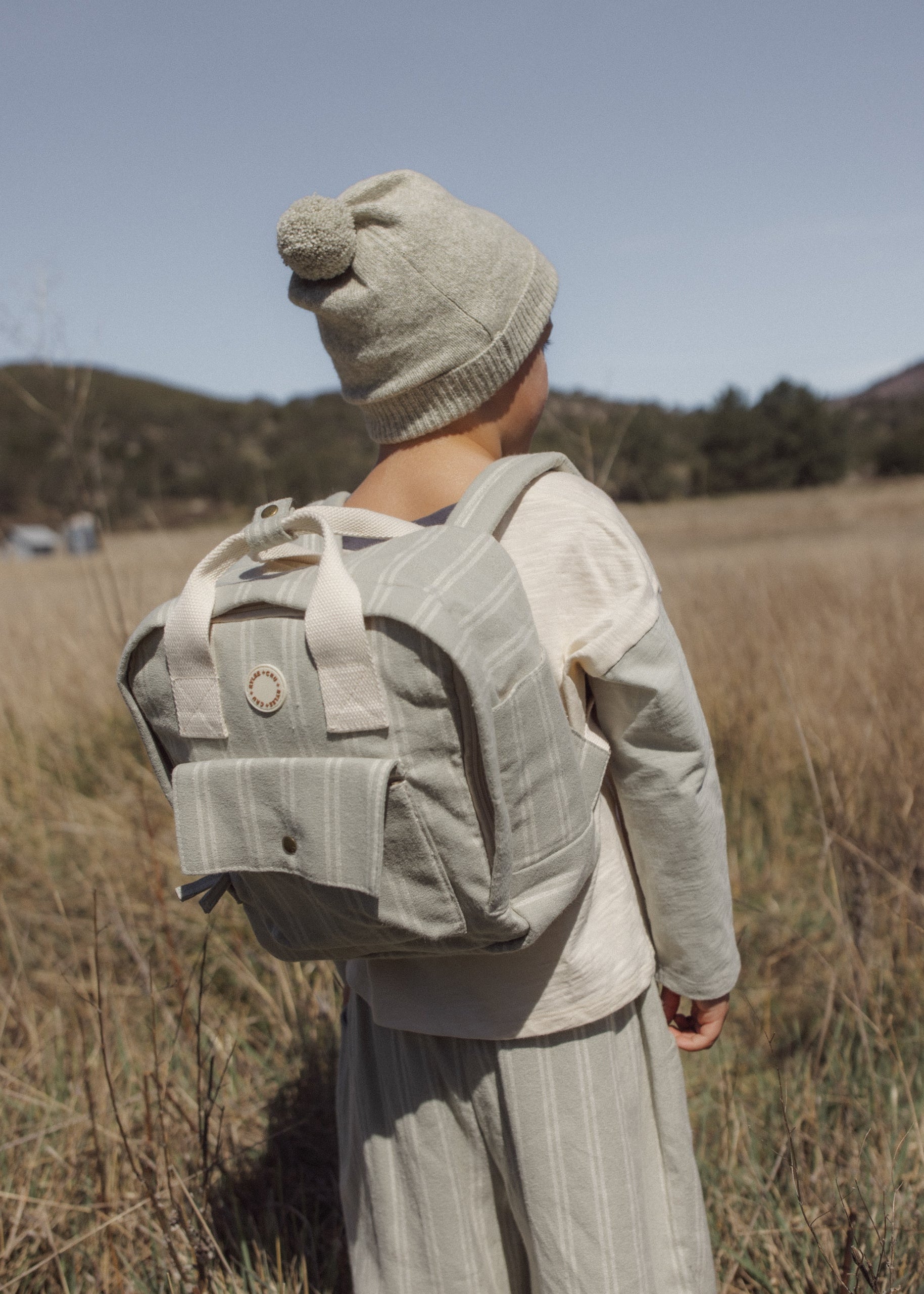 A child wearing a beige knit beanie and matching outfit stands in a dry grassy field, viewed from behind while wearing a light grayish-beige backpack.