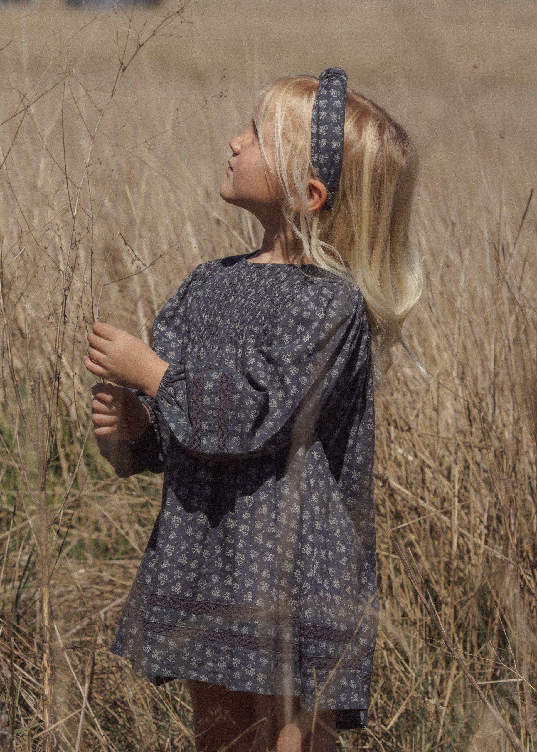 A young girl with blonde hair stands in a field of dry grass, wearing a loose, short-sleeved dark gray patterned dress and a matching patterned headband.