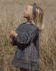 A young girl with blonde hair stands in a field of dry grass, wearing a loose, short-sleeved dark gray patterned dress and a matching patterned headband.