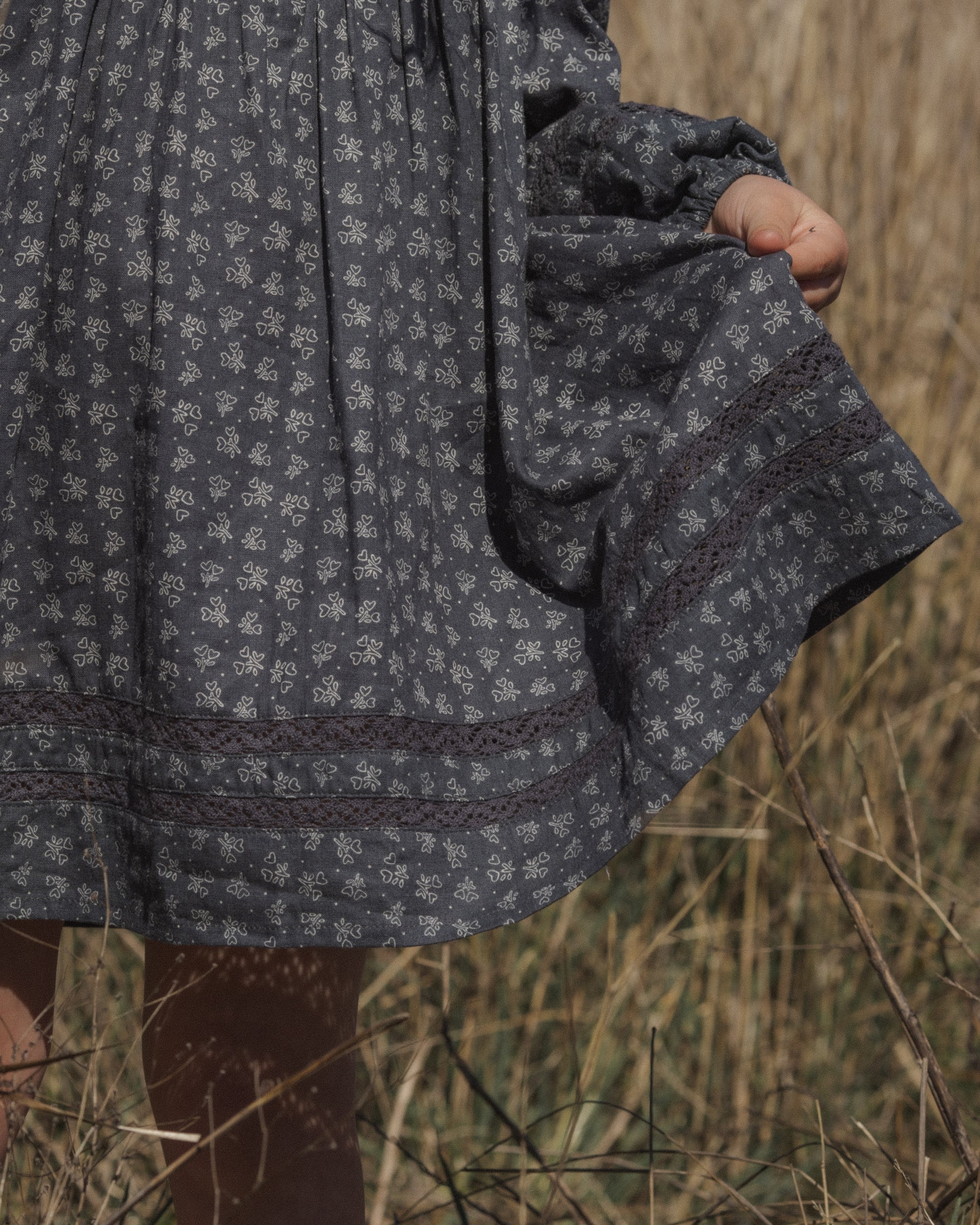 A close-up of a person outdoors wearing a dark patterned dress, holding a small brown bag, with dry grass in the background.