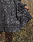 A close-up of a person outdoors wearing a dark patterned dress, holding a small brown bag, with dry grass in the background.
