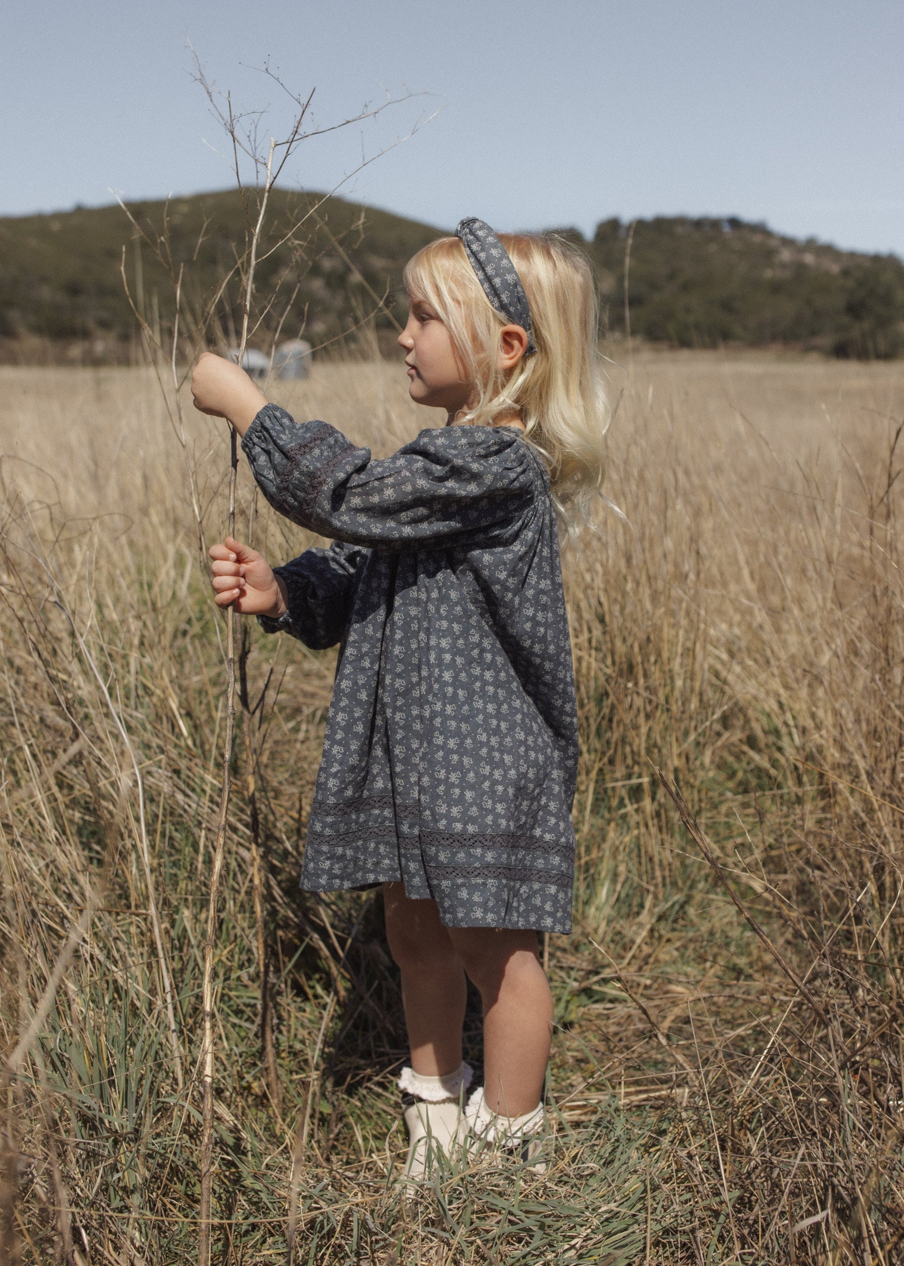 A young girl with blonde hair stands in a dry grassy field wearing a dark gray patterned long-sleeve dress, a matching headband, and white shoes.
