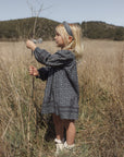 A young girl with blonde hair stands in a dry grassy field wearing a dark gray patterned long-sleeve dress, a matching headband, and white shoes.