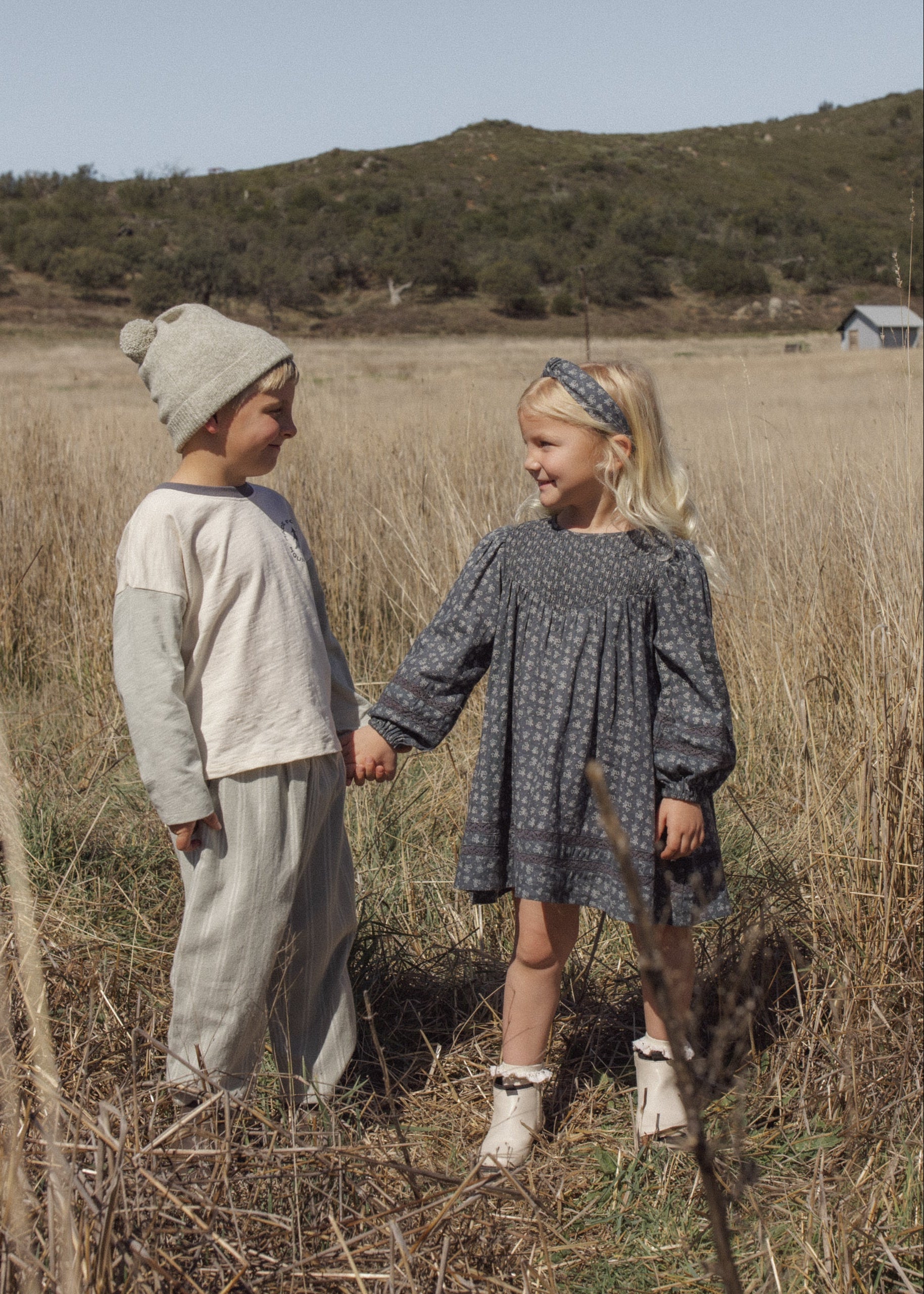 Two children standing in a dry grassy field outdoors, holding hands. The child on the left wears a light beige knit beanie, a light beige long-sleeve top, and light beige pants.