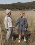 Two children standing in a dry grassy field outdoors, holding hands. The child on the left wears a light beige knit beanie, a light beige long-sleeve top, and light beige pants.