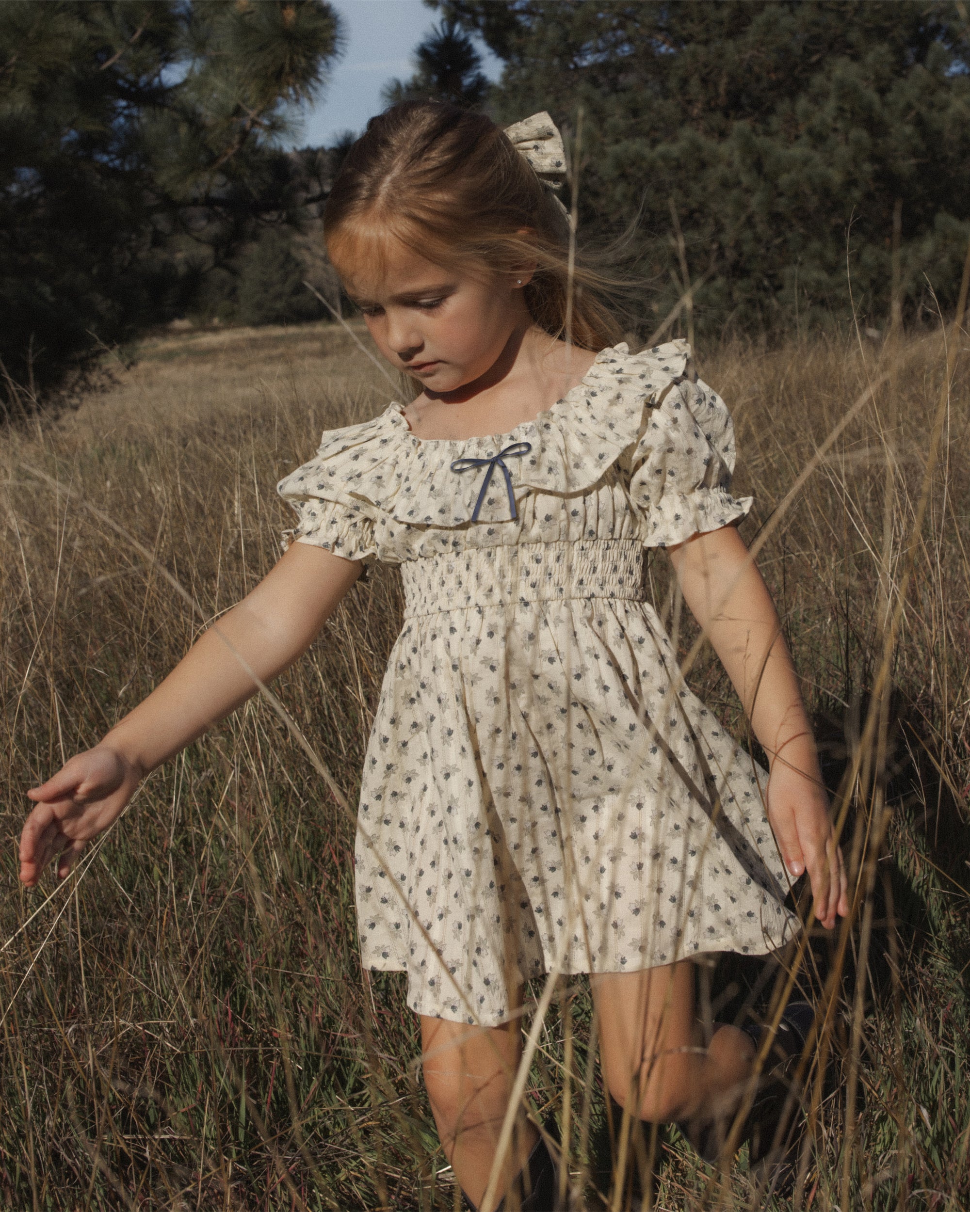 A young girl wearing a cream-colored short-sleeve dress with a small floral print and a blue bow at the neckline, walking through a dry grassy field outdoors.