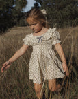 A young girl wearing a cream-colored short-sleeve dress with a small floral print and a blue bow at the neckline, walking through a dry grassy field outdoors.