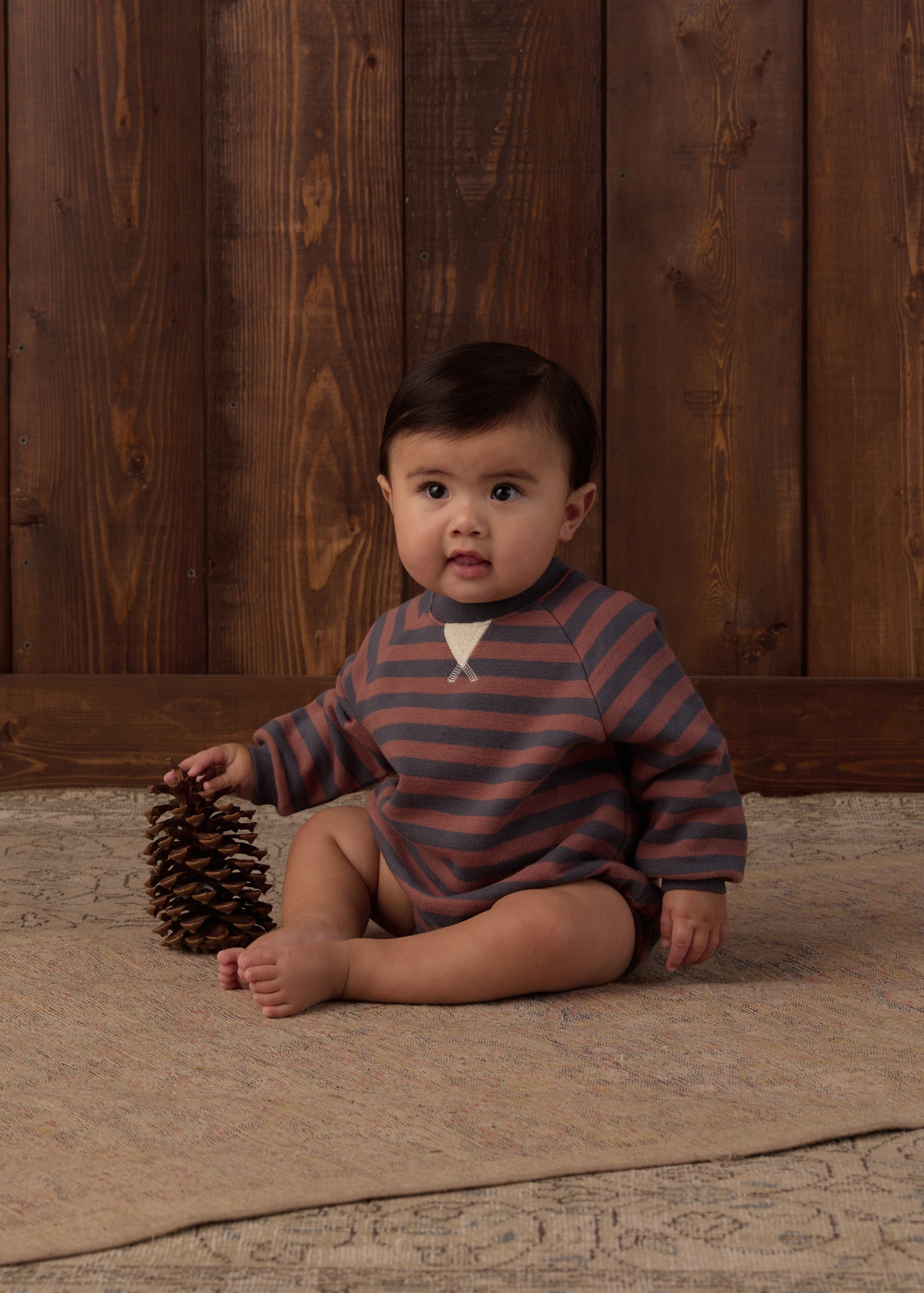 A baby sitting on a beige rug in front of a dark wood plank wall, wearing a long-sleeve striped romper in muted blue-gray and brown with a small white chest detail. The baby is holding a pinecone.