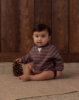A baby sitting on a beige rug in front of a dark wood plank wall, wearing a long-sleeve striped romper in muted blue-gray and brown with a small white chest detail. The baby is holding a pinecone.