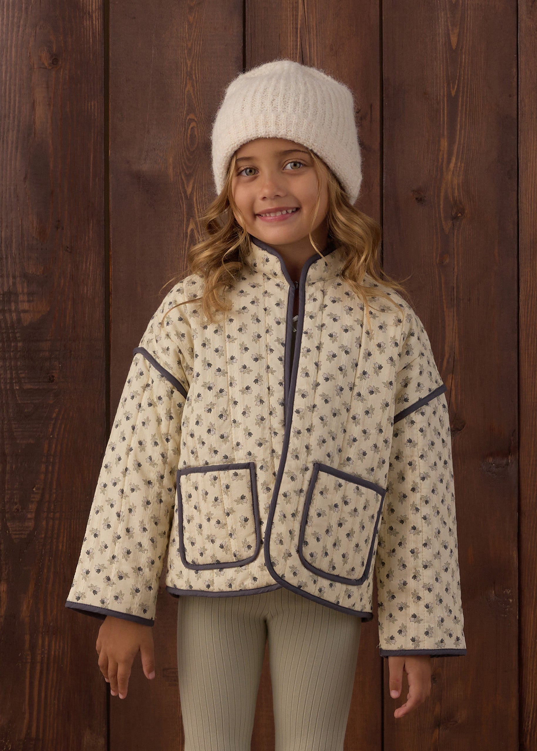 A young girl wearing a cream-colored fleece jacket with a small brown pattern and dark navy trim, a matching cream knit beanie, and ribbed cream leggings, standing against a dark brown wooden plank wall.