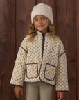 A young girl wearing a cream-colored fleece jacket with a small brown pattern and dark navy trim, a matching cream knit beanie, and ribbed cream leggings, standing against a dark brown wooden plank wall.