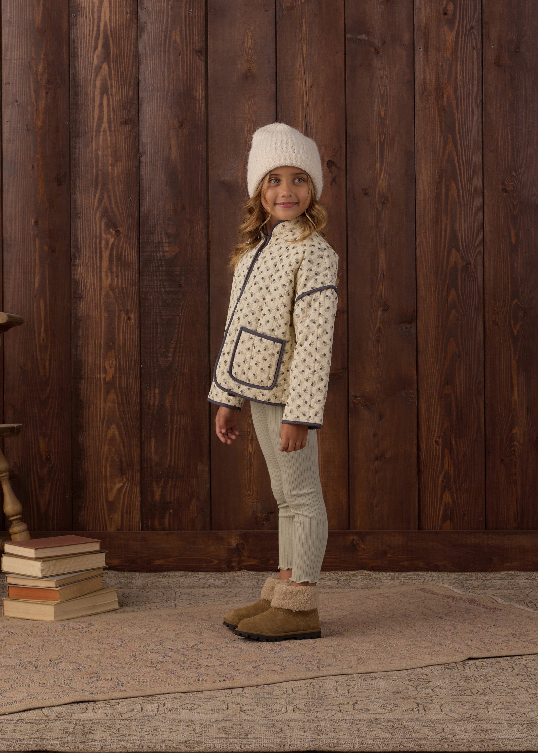 A young girl stands in front of a dark wood plank wall wearing a cream knit beanie, a cream patterned jacket with dark trim, light beige leggings, and tan slippers. A stack of books and part of a wooden chair are visible on the left.