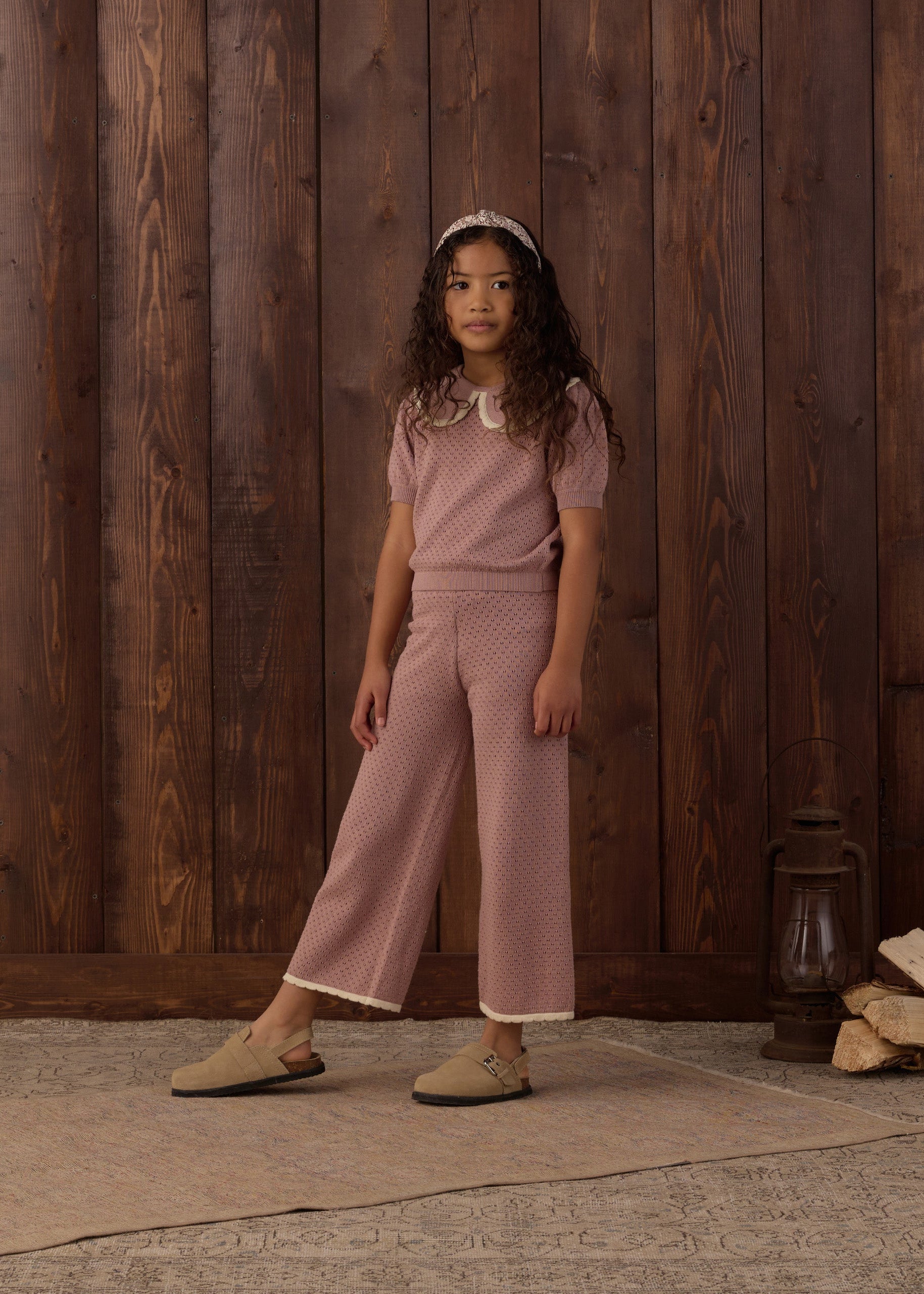 A young girl stands in front of a rustic dark wood plank wall wearing a dusty rose pink two-piece knit set consisting of a short-sleeve top with a white collar and matching wide-leg cropped pants. She is wearing beige slip-on shoes and a white headband.