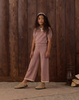 A young girl stands in front of a rustic dark wood plank wall wearing a dusty rose pink two-piece knit set consisting of a short-sleeve top with a white collar and matching wide-leg cropped pants. She is wearing beige slip-on shoes and a white headband.