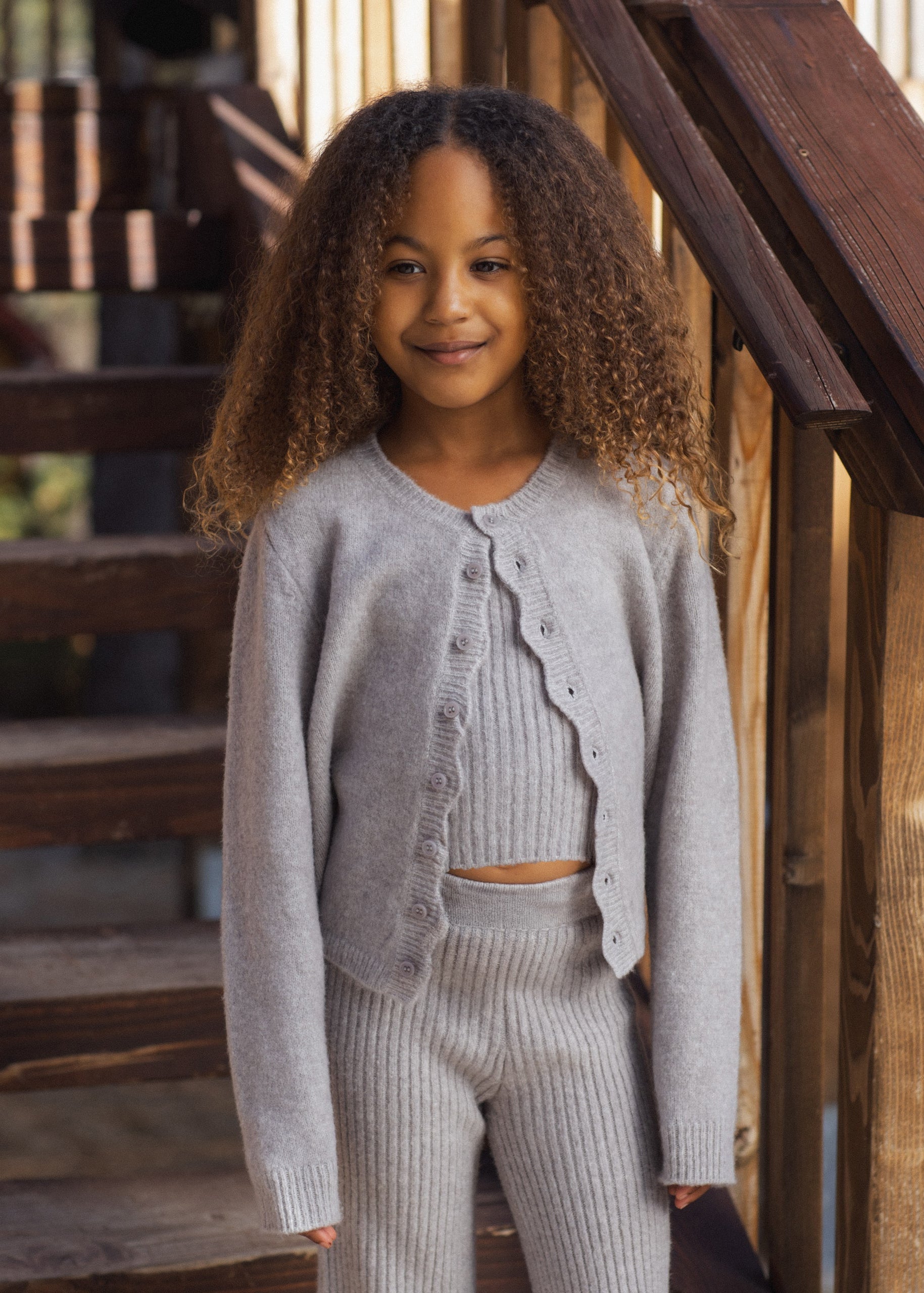 A young girl with curly hair stands on wooden stairs wearing a light gray knit cardigan and matching ribbed pants.