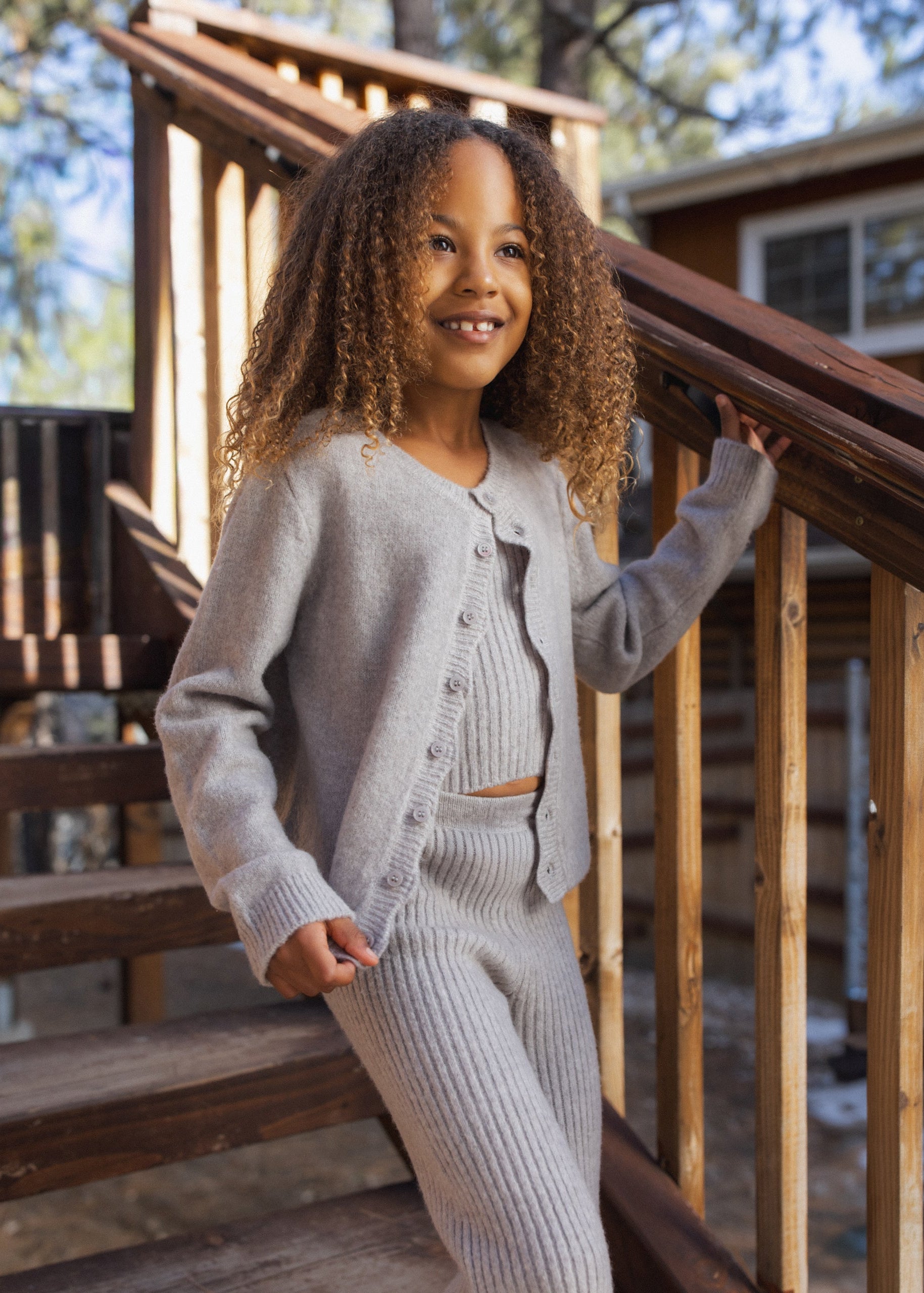 A young girl with curly hair stands on wooden outdoor stairs wearing a light gray ribbed knit cardigan and matching pants set. She is smiling and posing next to a wooden railing in front of a cabin background.