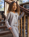A young girl with curly hair stands on wooden outdoor stairs wearing a light gray ribbed knit cardigan and matching pants set. She is smiling and posing next to a wooden railing in front of a cabin background.