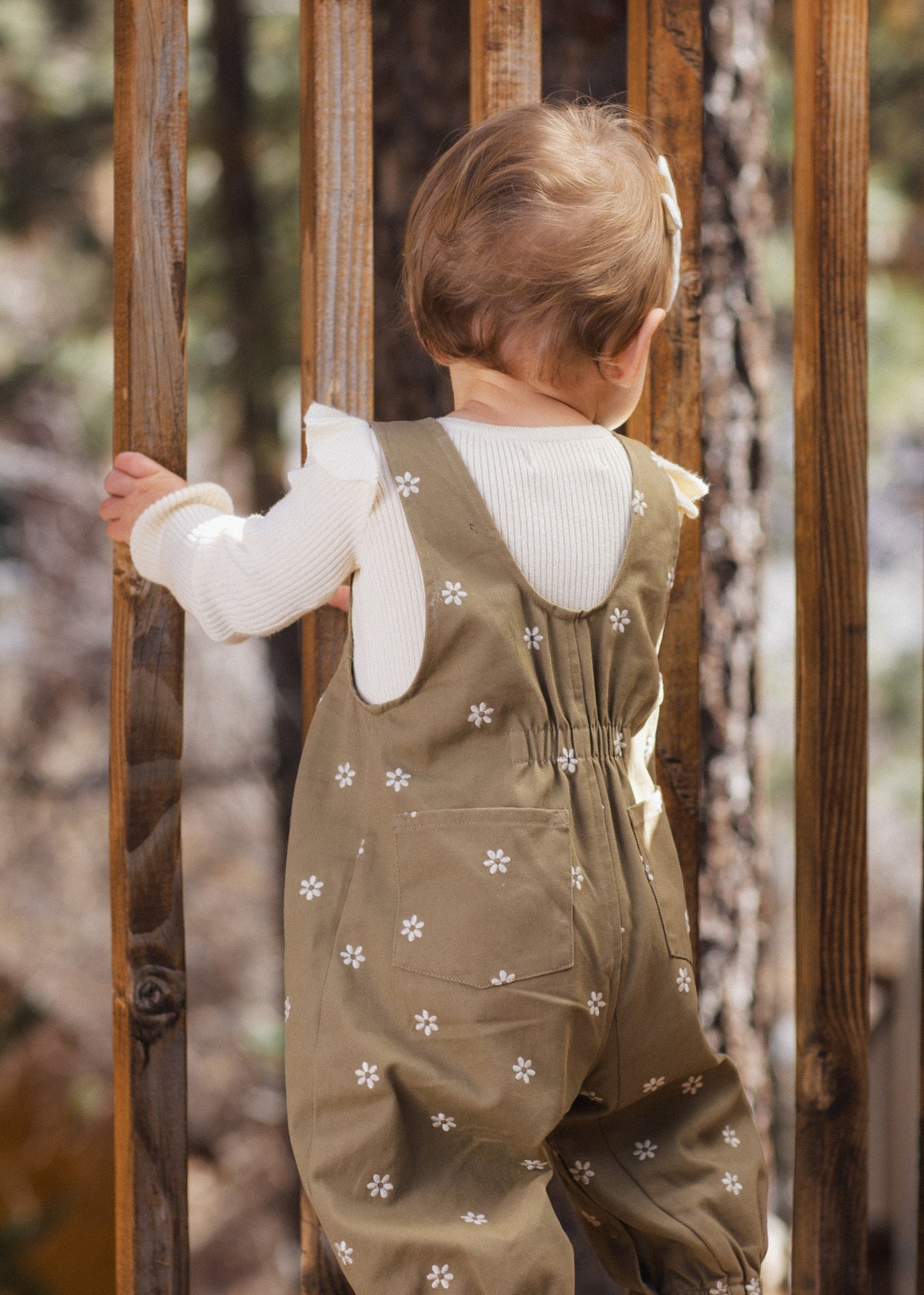 A baby wearing olive green floral overalls and a white ribbed long-sleeve top, seen from behind while climbing over a wooden fence outdoors.