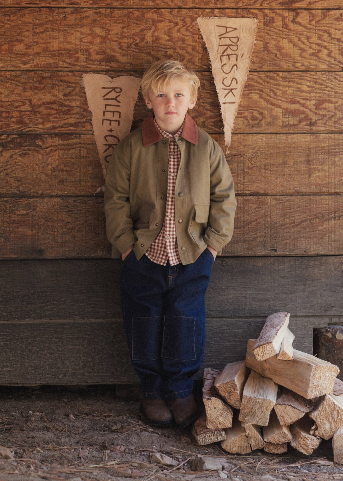 A young boy stands against a rustic wooden wall wearing a brown jacket, red-and-white check shirt, and dark blue jeans, with a stack of firewood beside him and two pennant flags behind him.