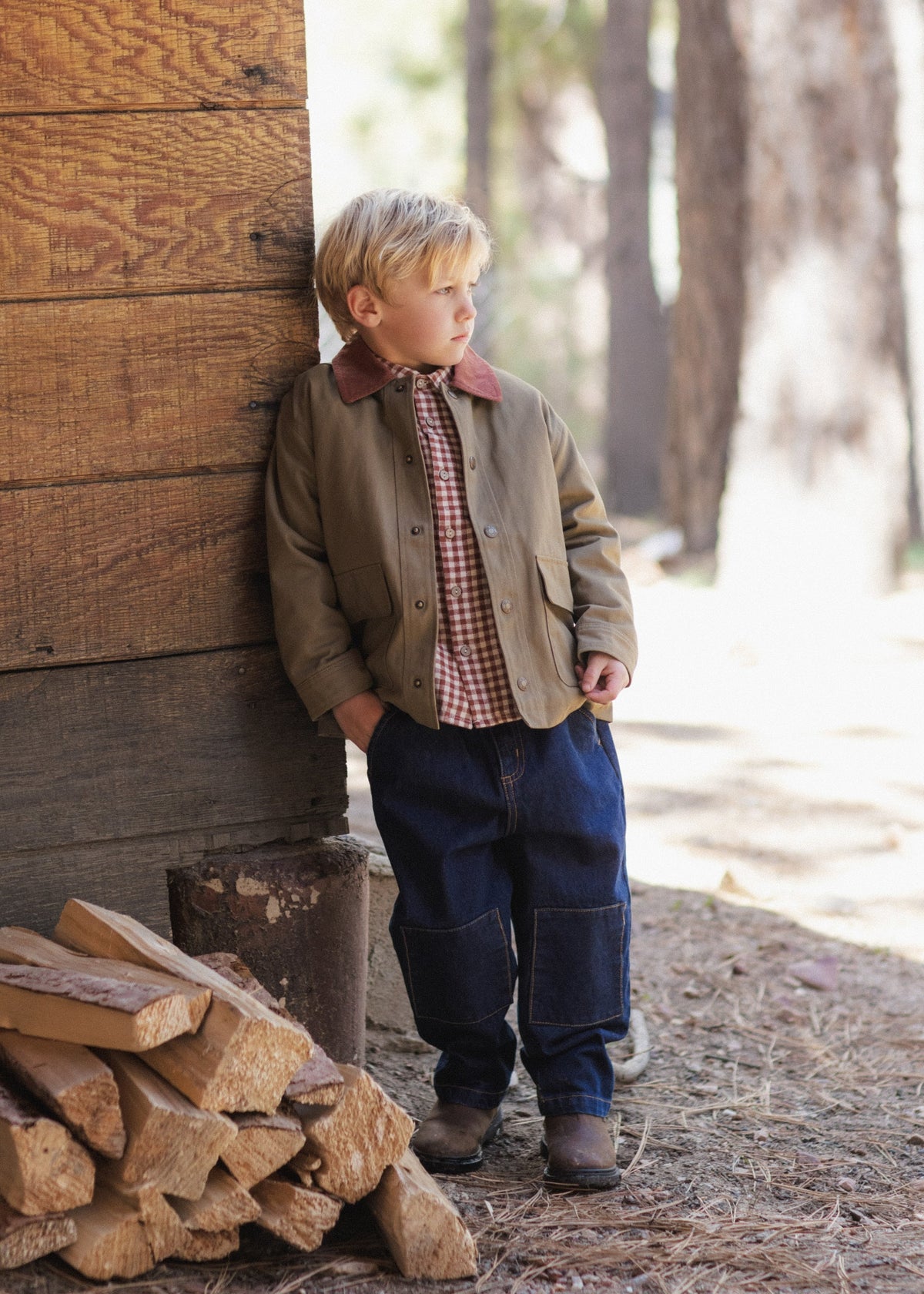 A young boy stands outdoors leaning against a rustic wooden wall next to a stack of firewood. He is wearing a brown corduroy jacket with a reddish-brown collar, a red-and-white gingham shirt, dark blue jeans, and brown boots.