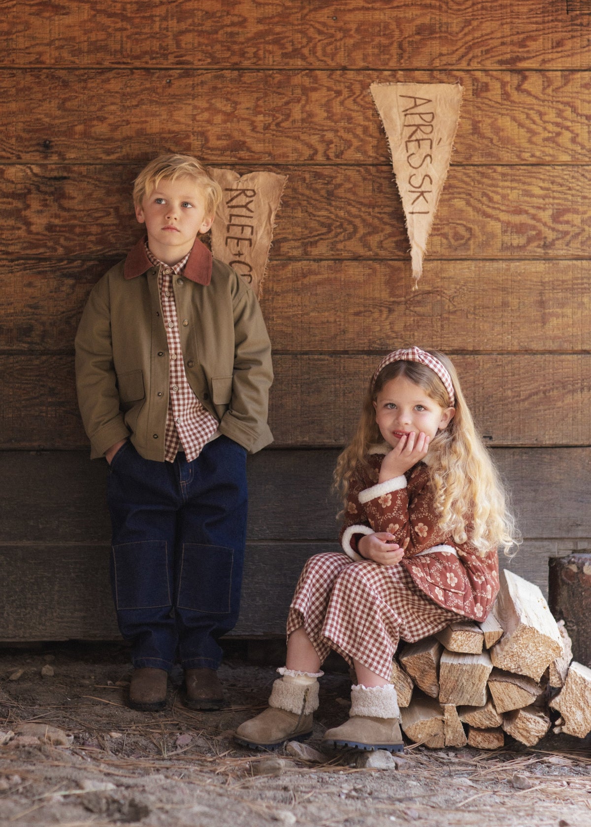 Two children posing outdoors against a rustic wooden wall. The boy stands on the left wearing a brown jacket, red-and-white gingham shirt and tie, and dark blue jeans.
