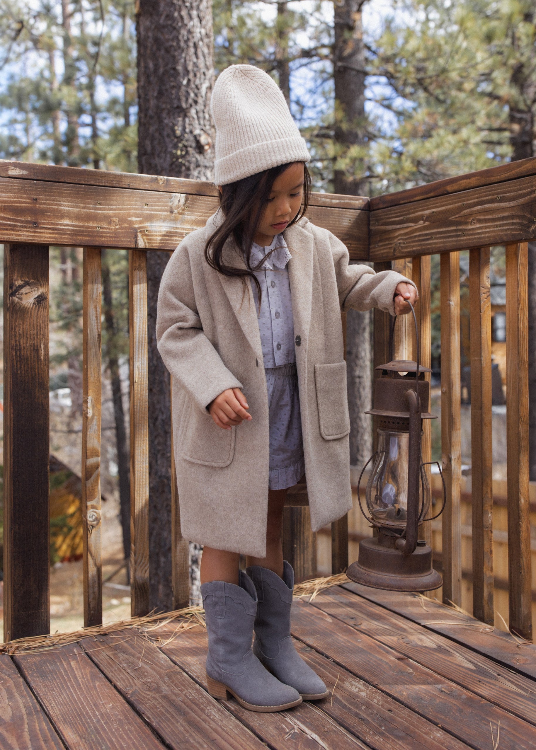 A young girl stands on a wooden deck wearing a long beige coat, a cream knit beanie, a blue button-up shirt, a gray skirt, and gray boots, while holding a vintage-style lantern. Trees and a cabin are visible in the background.