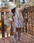 A young girl stands on a wooden deck wearing a beige knit beanie, a light beige oversized coat, a light blue patterned crop top and shorts set, and gray cowboy boots while holding a vintage-style lantern.