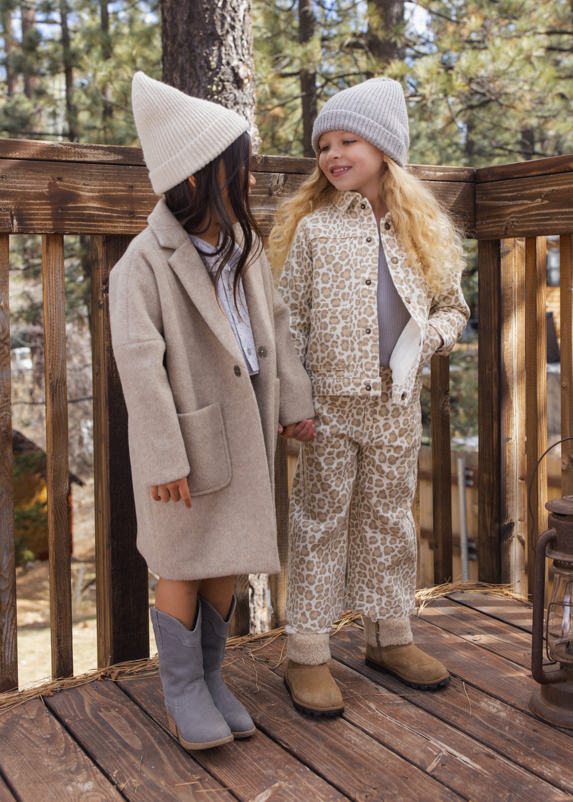 Two young girls standing on a wooden deck outdoors, wearing coordinated fall fashion. One girl wears a beige coat with a matching beanie and gray boots, while the other wears a beige-and-white animal-print jacket and matching pants with a light knit beanie and tan boots.