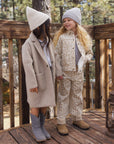 Two young girls standing on a wooden deck outdoors, wearing coordinated fall fashion. One girl wears a beige coat with a matching beanie and gray boots, while the other wears a beige-and-white animal-print jacket and matching pants with a light knit beanie and tan boots.