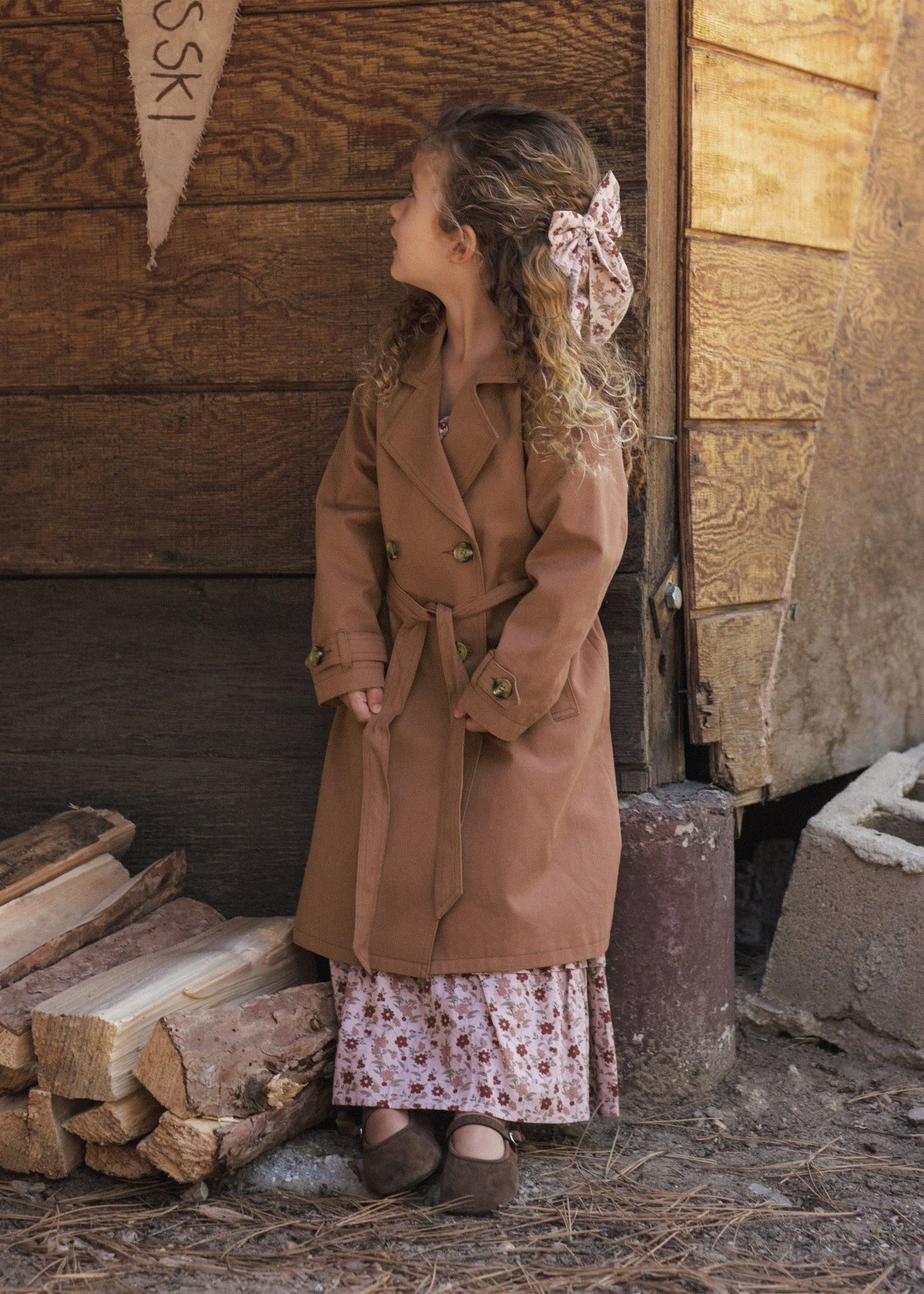 A young girl with curly hair wearing a brown belted trench coat over a pink floral dress, standing outdoors in front of a rustic wooden wall next to a stack of firewood.