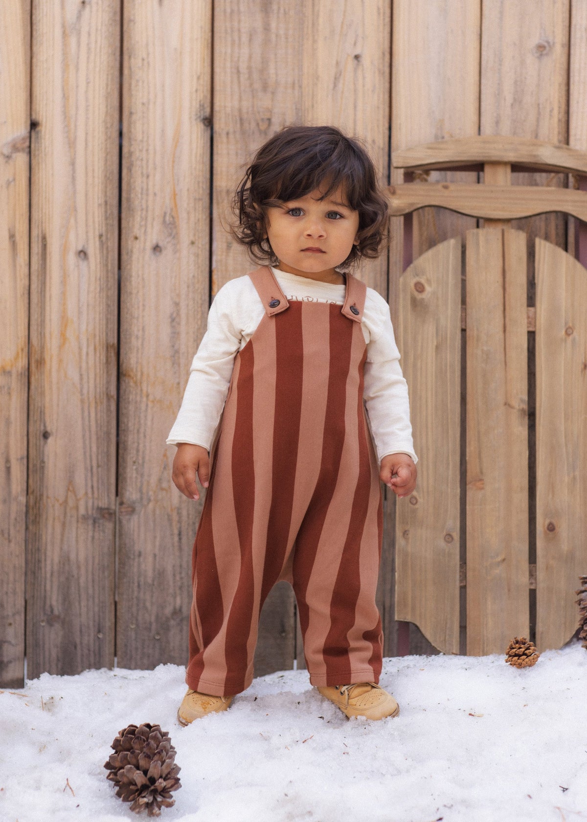 A toddler stands in snow in front of a wooden fence, wearing rust and tan vertically striped overalls over a cream long-sleeve shirt and tan boots.