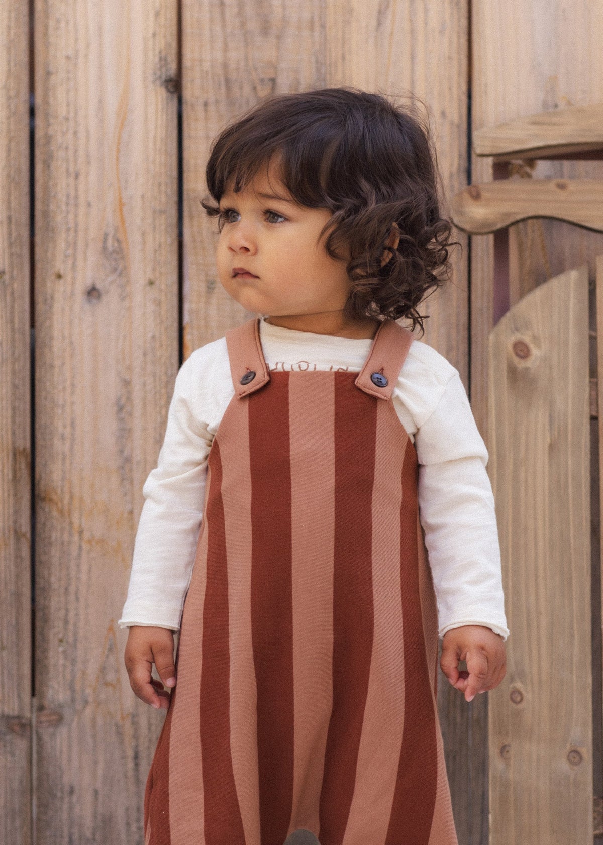 A toddler with dark curly hair wearing a white long-sleeve shirt and a rust-brown and dusty pink vertically striped pinafore overalls, standing in front of a rustic wooden fence.