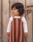 A toddler with dark curly hair wearing a white long-sleeve shirt and a rust-brown and dusty pink vertically striped pinafore overalls, standing in front of a rustic wooden fence.