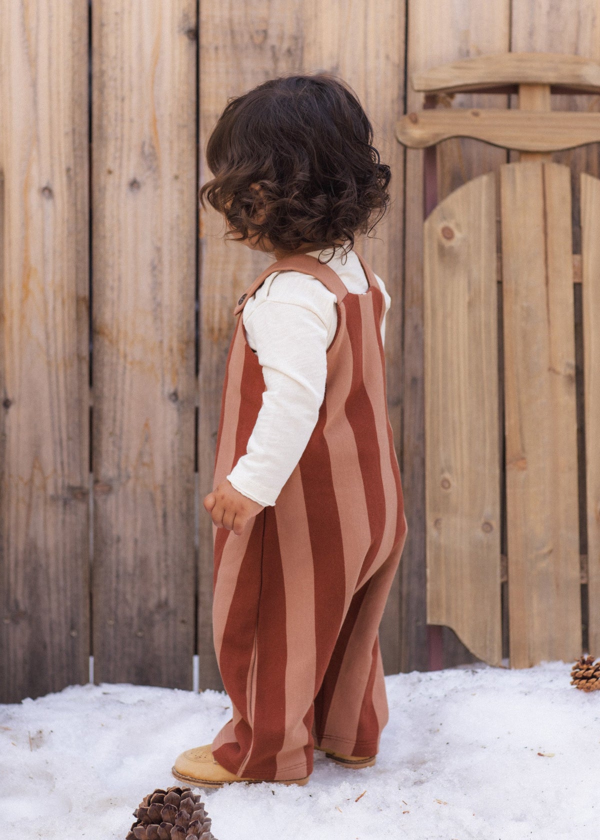 A toddler standing outdoors in front of a wooden fence and snow, wearing a rust and dusty pink vertically striped overall jumpsuit over a white long-sleeve shirt.