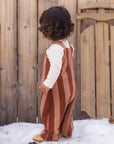 A toddler standing outdoors in front of a wooden fence and snow, wearing a rust and dusty pink vertically striped overall jumpsuit over a white long-sleeve shirt.