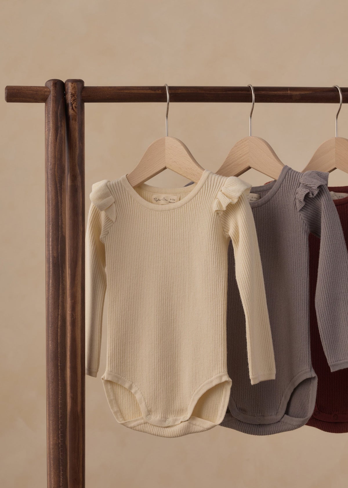 Three ribbed long-sleeve baby bodysuits hang on wooden hangers from a dark wood clothing rack against a warm beige background. The bodysuits are in cream, dusty gray, and burgundy.