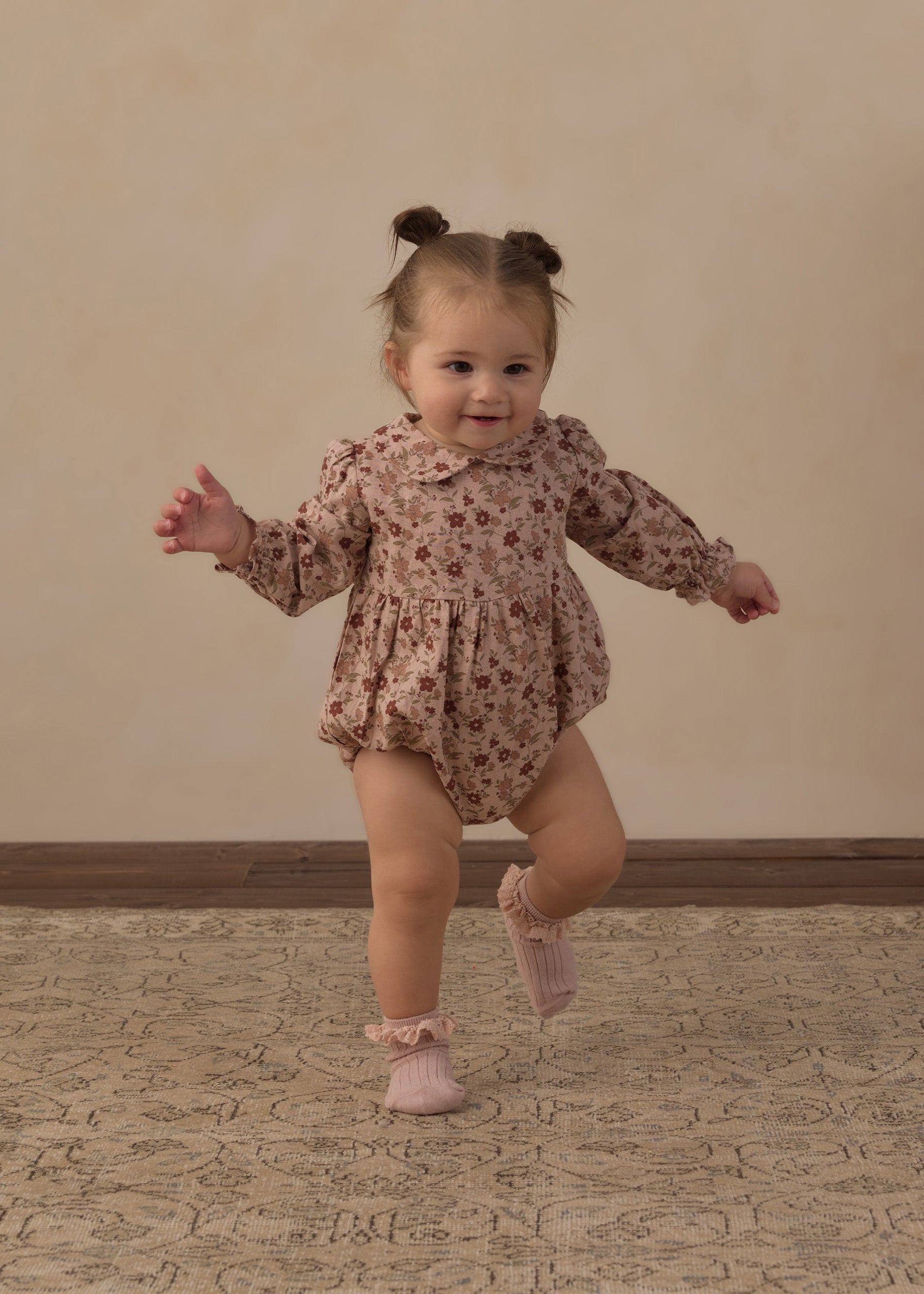 A toddler girl wearing a long-sleeve floral romper and pink booties is captured mid-step while running on a patterned rug against a beige wall.