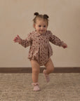 A toddler girl wearing a long-sleeve floral romper and pink booties is captured mid-step while running on a patterned rug against a beige wall.