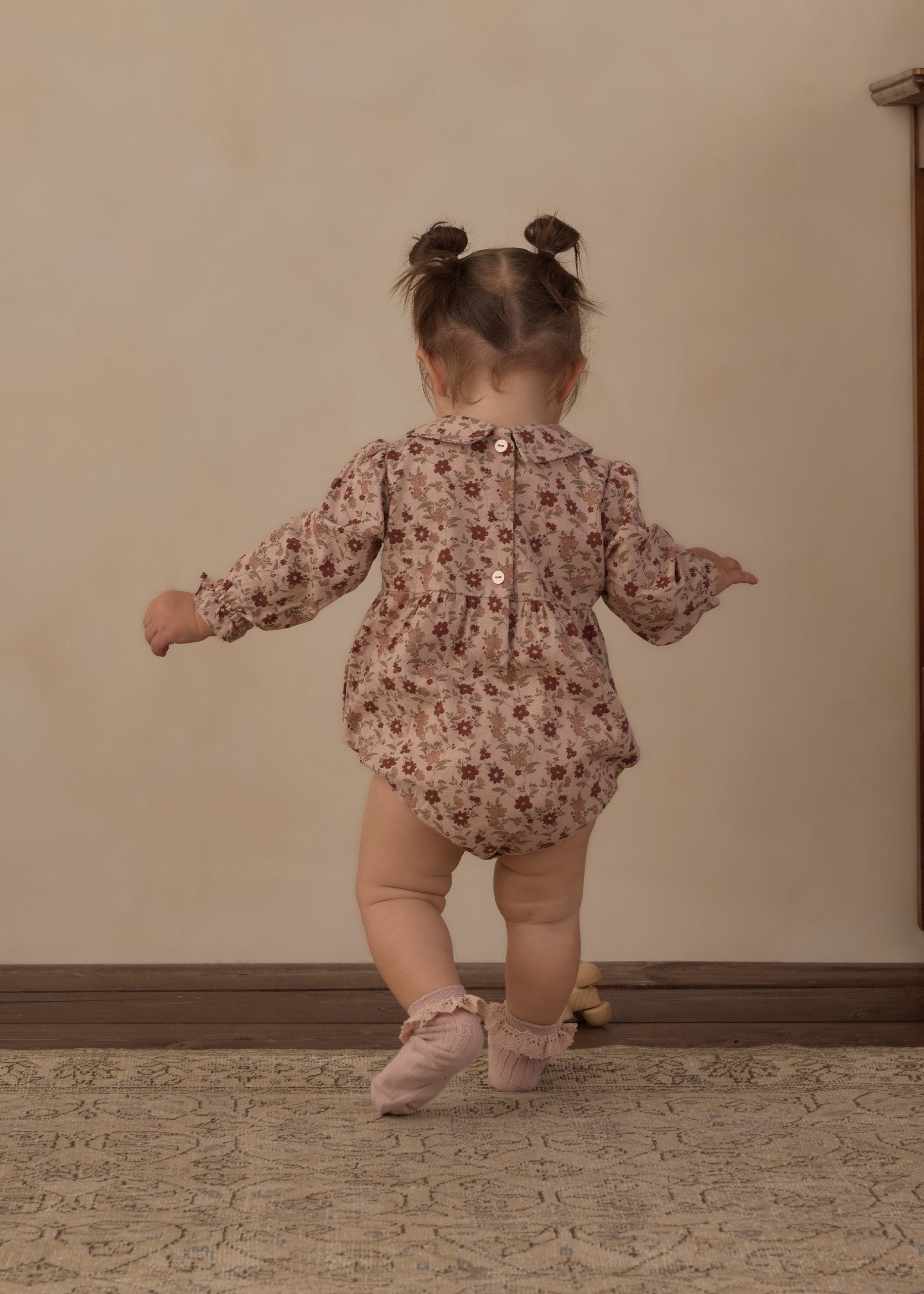 A toddler girl seen from behind wearing a long-sleeve floral romper and pink ballet shoes, standing on a patterned rug against a beige wall.