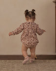 A toddler girl seen from behind wearing a long-sleeve floral romper and pink ballet shoes, standing on a patterned rug against a beige wall.