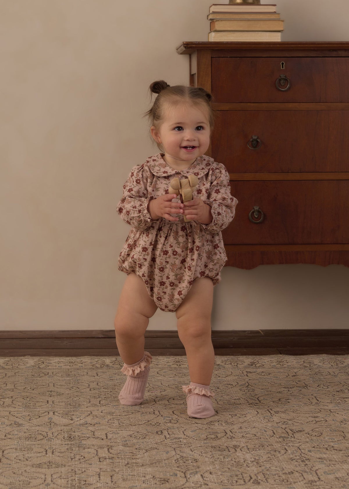 A toddler girl wearing a long-sleeve floral romper and pink booties is standing and smiling in a room with a wooden dresser and beige wall.