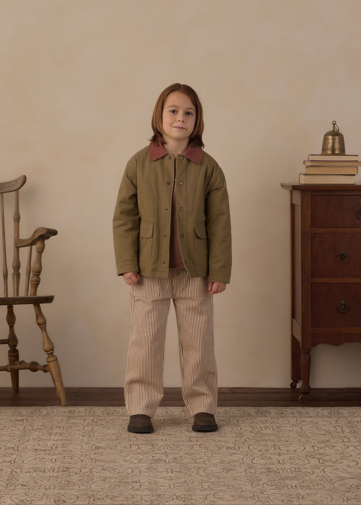 A young child stands in a room wearing a muted olive green jacket with brown collar panels, beige corduroy trousers, and dark shoes. The room has a textured beige wall, a wooden chair on the left, and a wooden dresser with books and a small brass bell on the right.