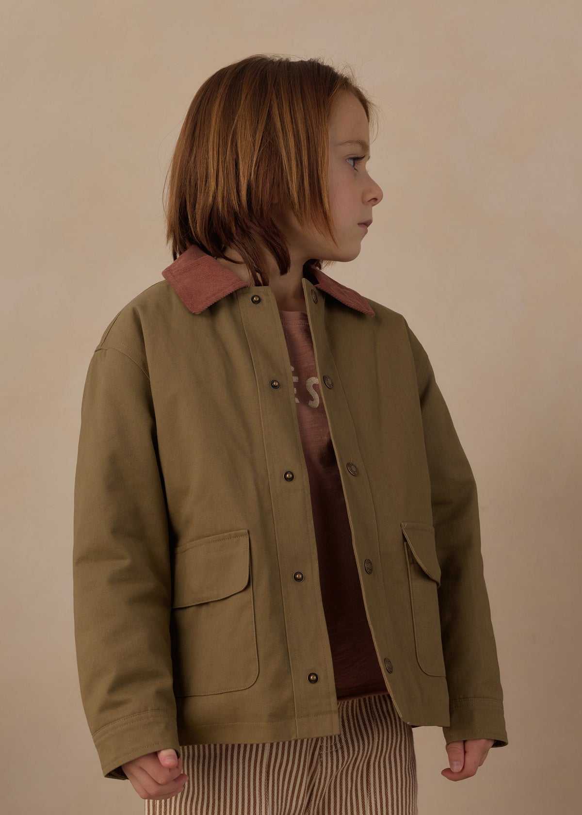 A child wearing an olive green jacket with a brown collar, layered over a brown top and striped pants, standing against a plain beige background.