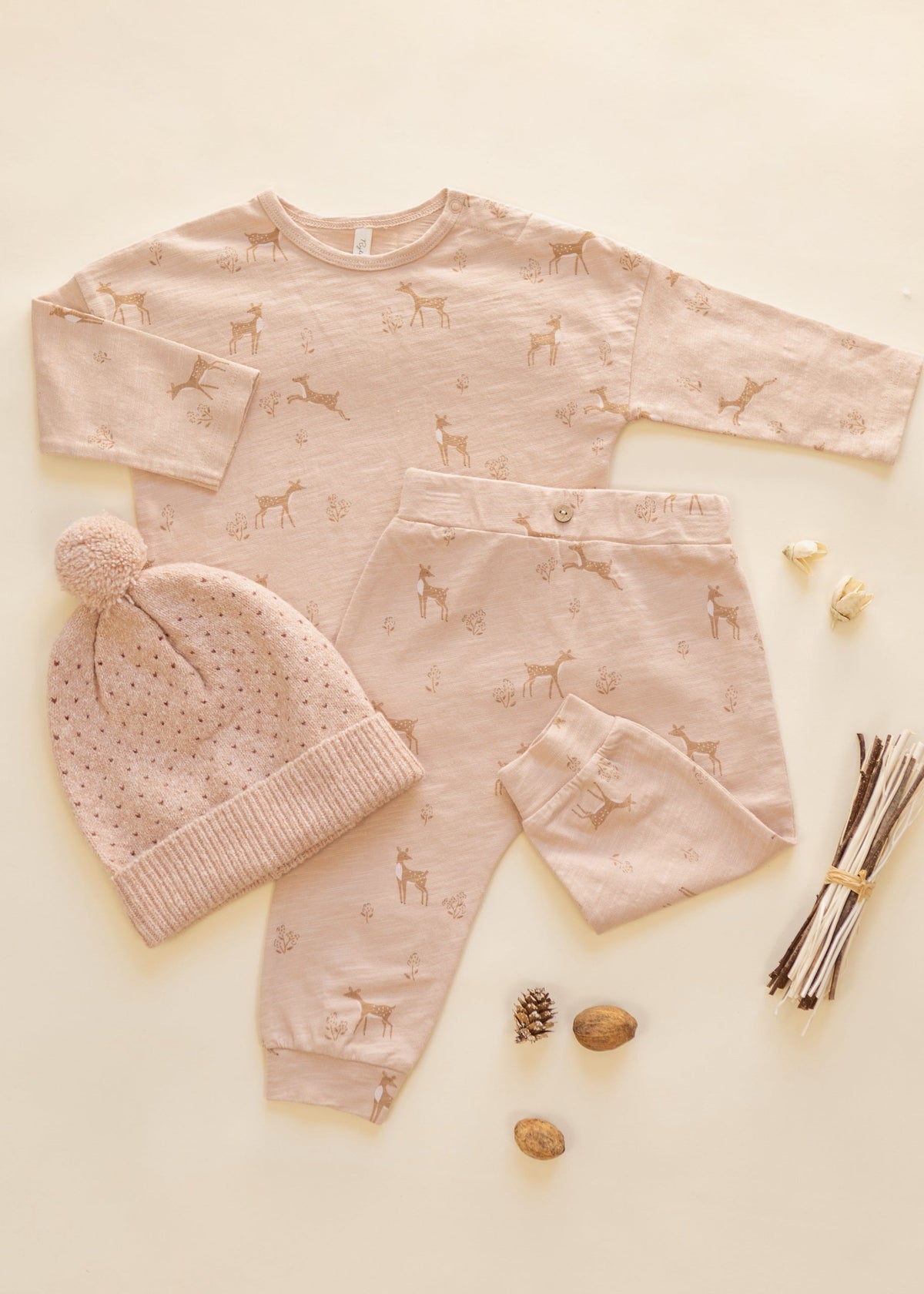 A baby clothing set laid flat on a warm cream background, including a long-sleeve top, pants, and a knit beanie with a pom-pom. The set is light blush pink with a subtle deer print and small polka dots, styled with natural props like pinecones, acorns, and twigs.